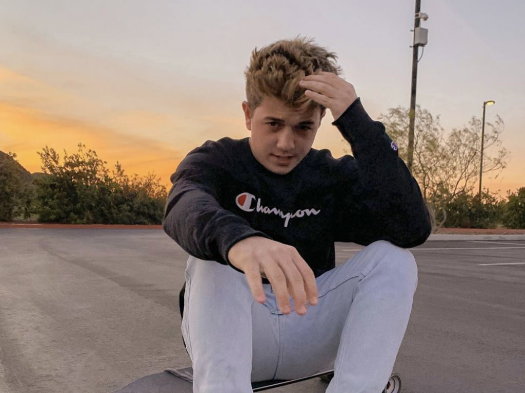 Bradley Steven Perry sitting on a skateboard in a parking lot.