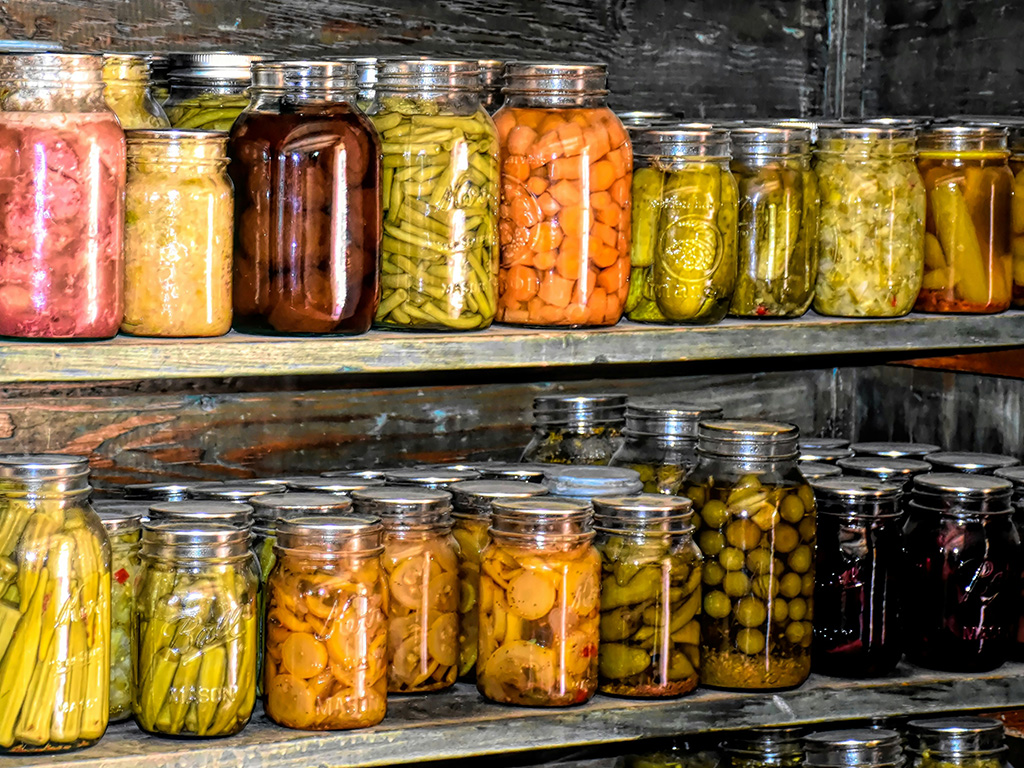 Two shelves of canned food including green beans, olives and cucumbers. 