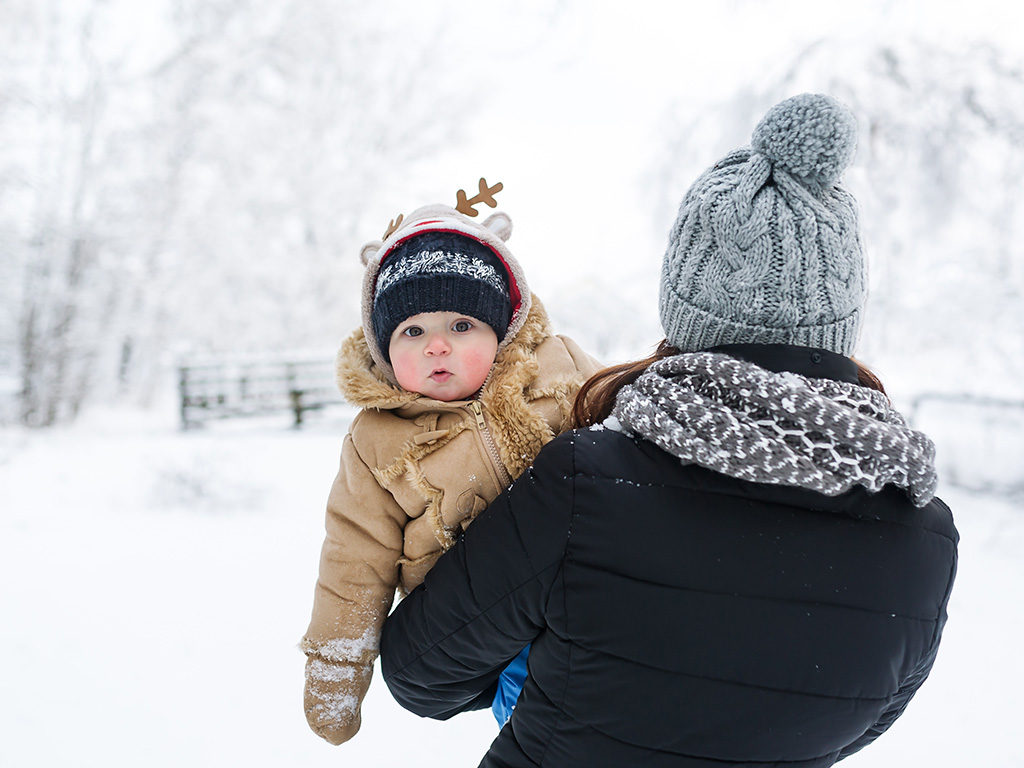 A boy son looks out from behind mother. The mother carries the baby in winter park. Closeup portrait