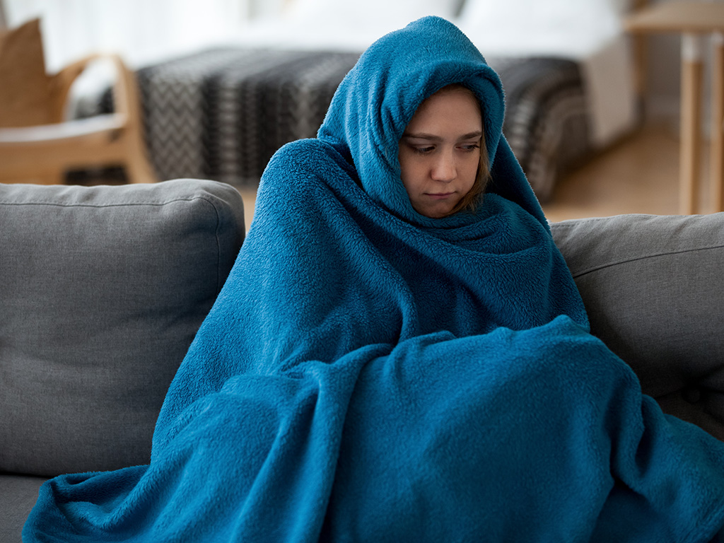 Young unsatisfied sick woman covered with warm blue blanket plaid from head to toes sitting alone on couch in living room at cold home. 