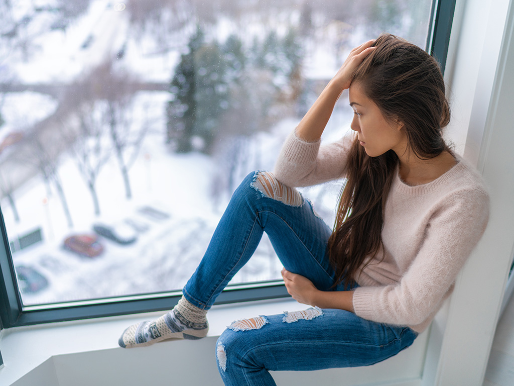 Winter depressed sad girl lonely by home window looking at cold weather upset unhappy.