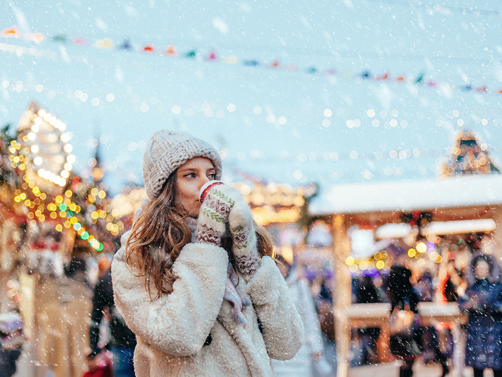 Girl drinking hot coffee while walking in Christmas market decorated with holiday lights in the evening. 