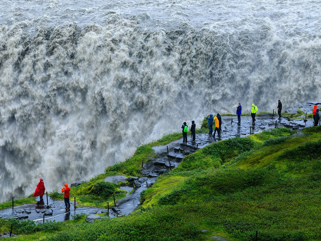 Tourists at a wet and powerful Dettifoss waterfall as it gushes into Jökulsárgljúfur canyon, with rising mist and spray and green Summer grasses, Vatnajökull National Park, North Iceland, Europe