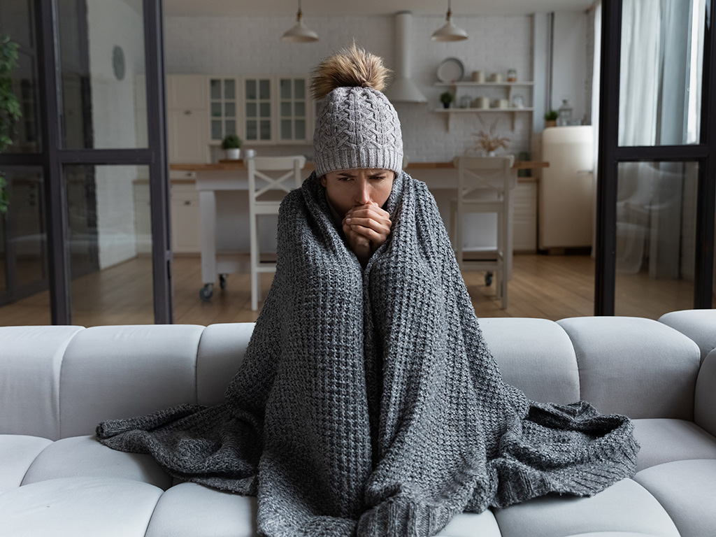 Person sitting on the couch with a blanket wrapped around them while wearing a hat