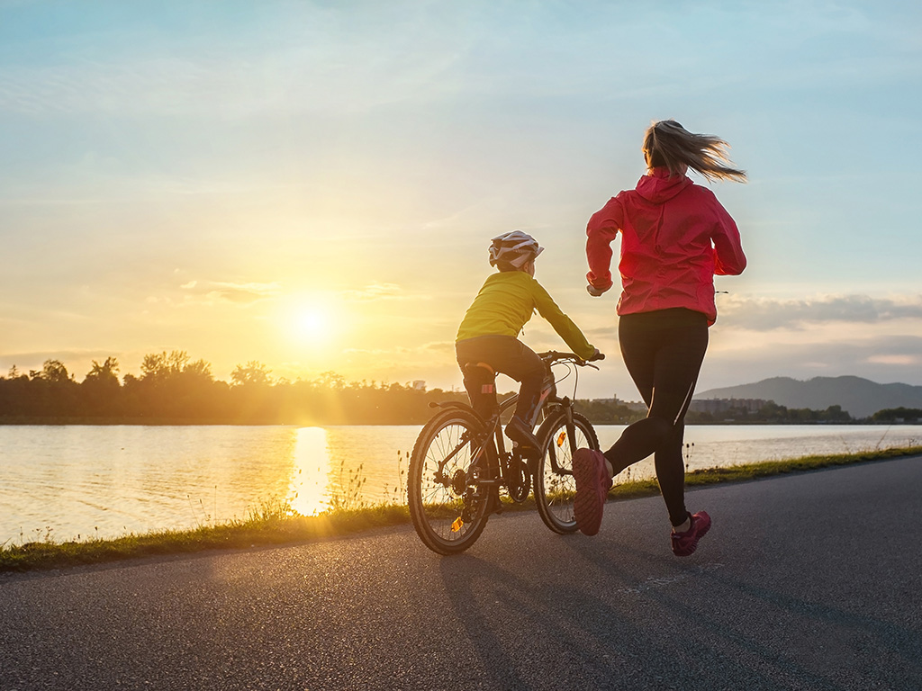 Boy rides bike in helmets, mom runs on sunny day. 