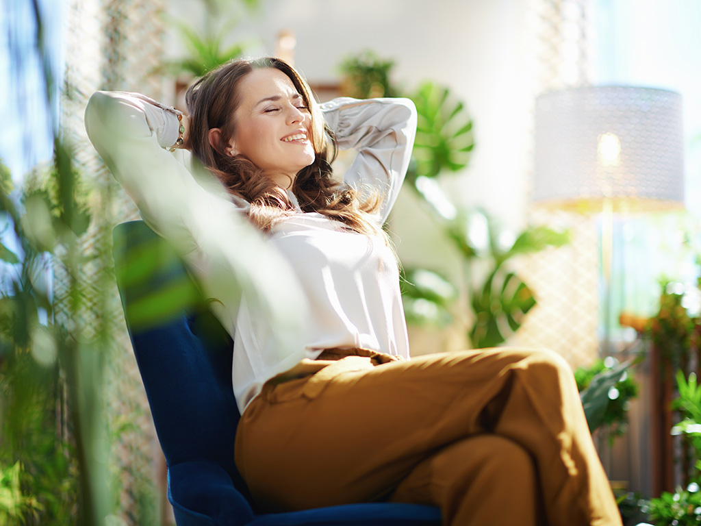 Relaxed stylish woman with long wavy hair at modern home in sunny day in green pants and grey blouse sitting in a blue armchair.