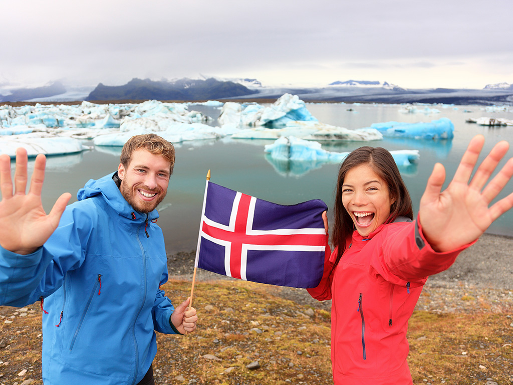 Tourist couple happy holding the Icelandic flag in front of the glacial lake / glacier lagoon.