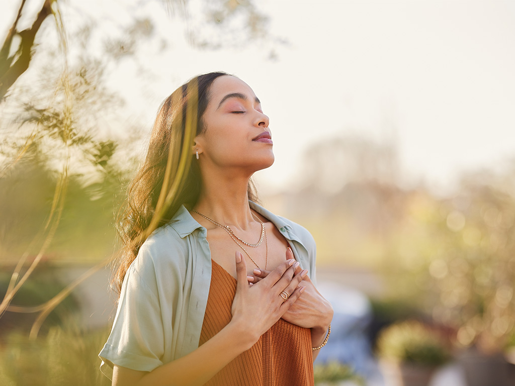 Young latin woman with hand on chest breathing in fresh air in a beautiful garden during sunset. 