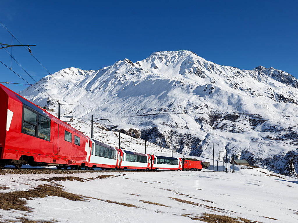 On a sunny winter day, tourists ride in a Glacier Express train and, thru the panoramic windows, enjoy the view of snowy Rossbodenstock mountain under blue clear sky, in Andermatt, Uri, Switzerland