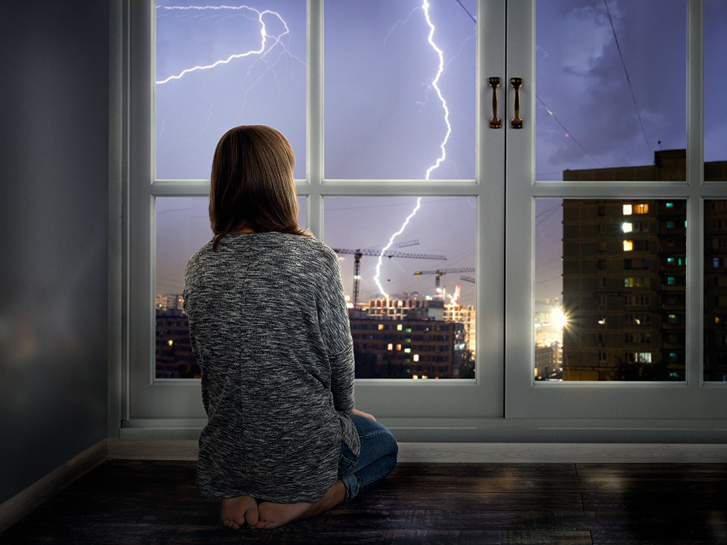 The girl looks through the window at the lightning. 