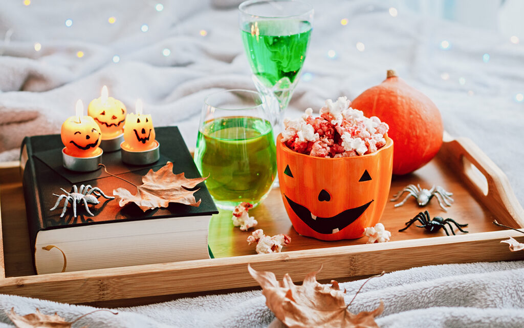 Book, halloween decor, candles, popcorn and drinks on tray on bed