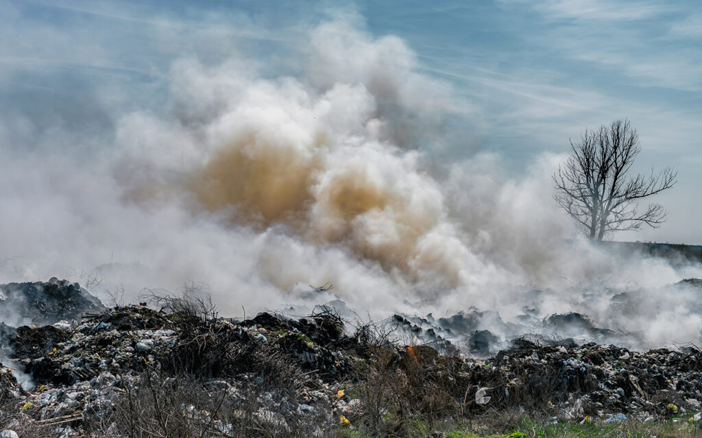 Fire at landfill with white smoke in sunny weather and clouded sky.